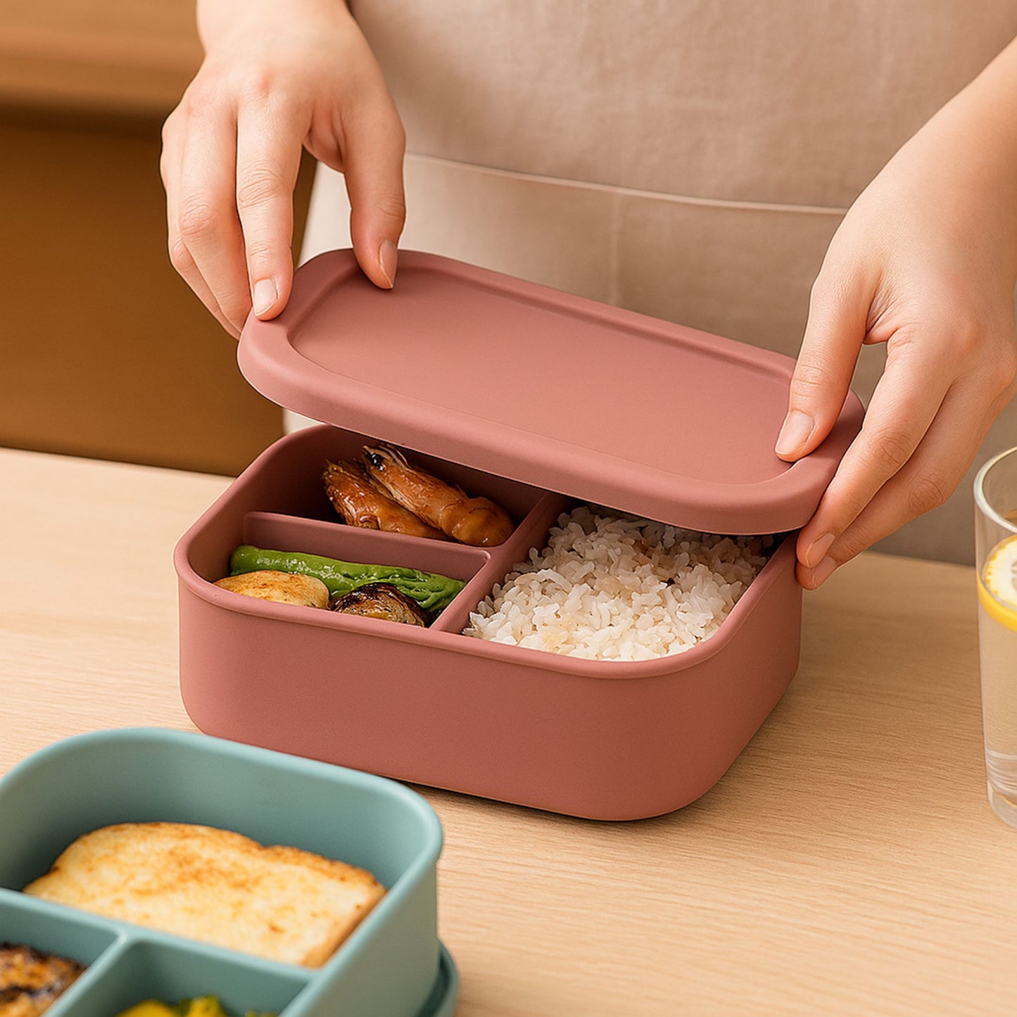 A person closing a pink silicone bento lunchbox filled with rice, shrimp, and sautéed vegetables. A drink and another blue lunchbox are visible in the background on a wooden table