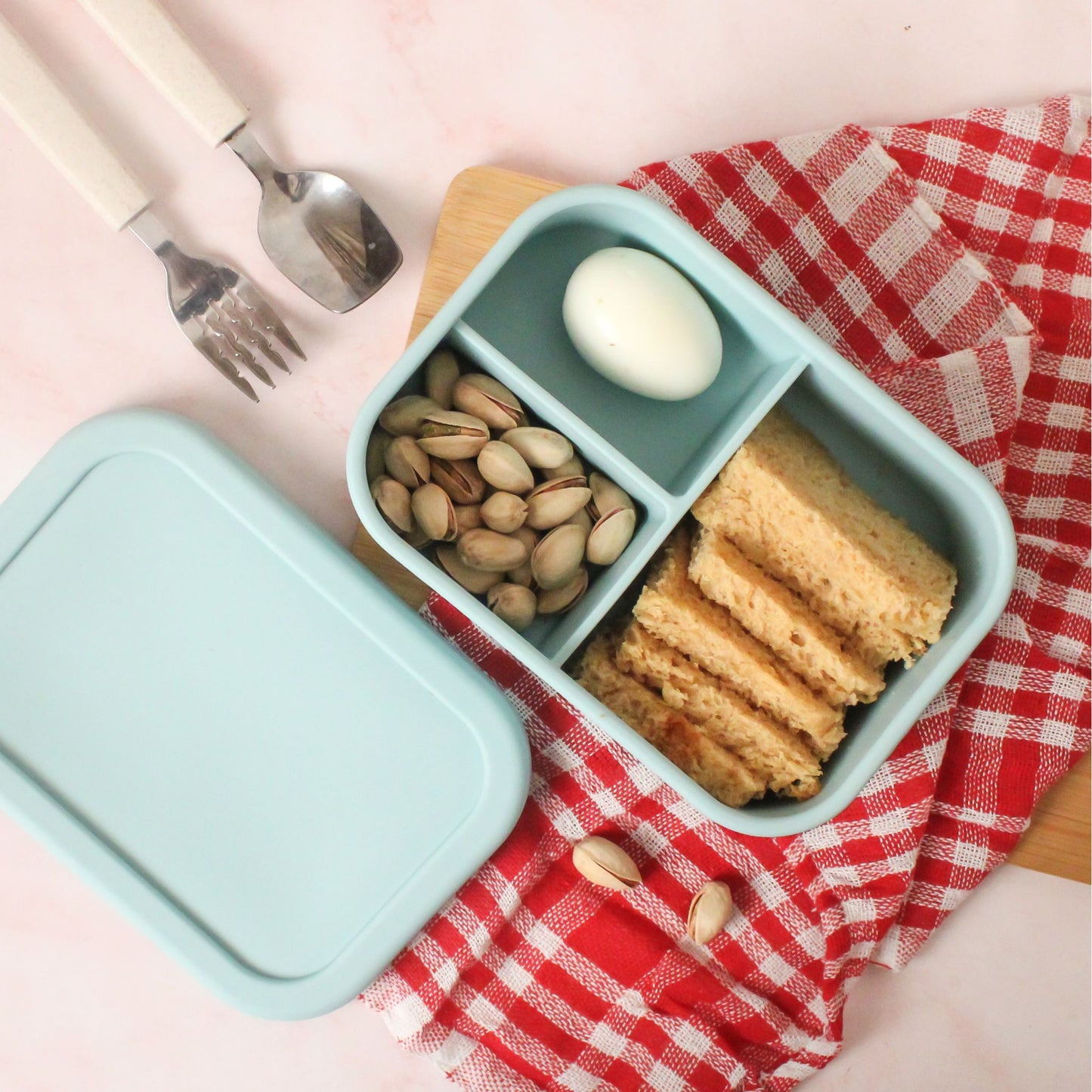 A blue silicone lunchbox containing bread slices, boiled egg, and pistachios placed on a red-checkered napkin for a breakfast setup.