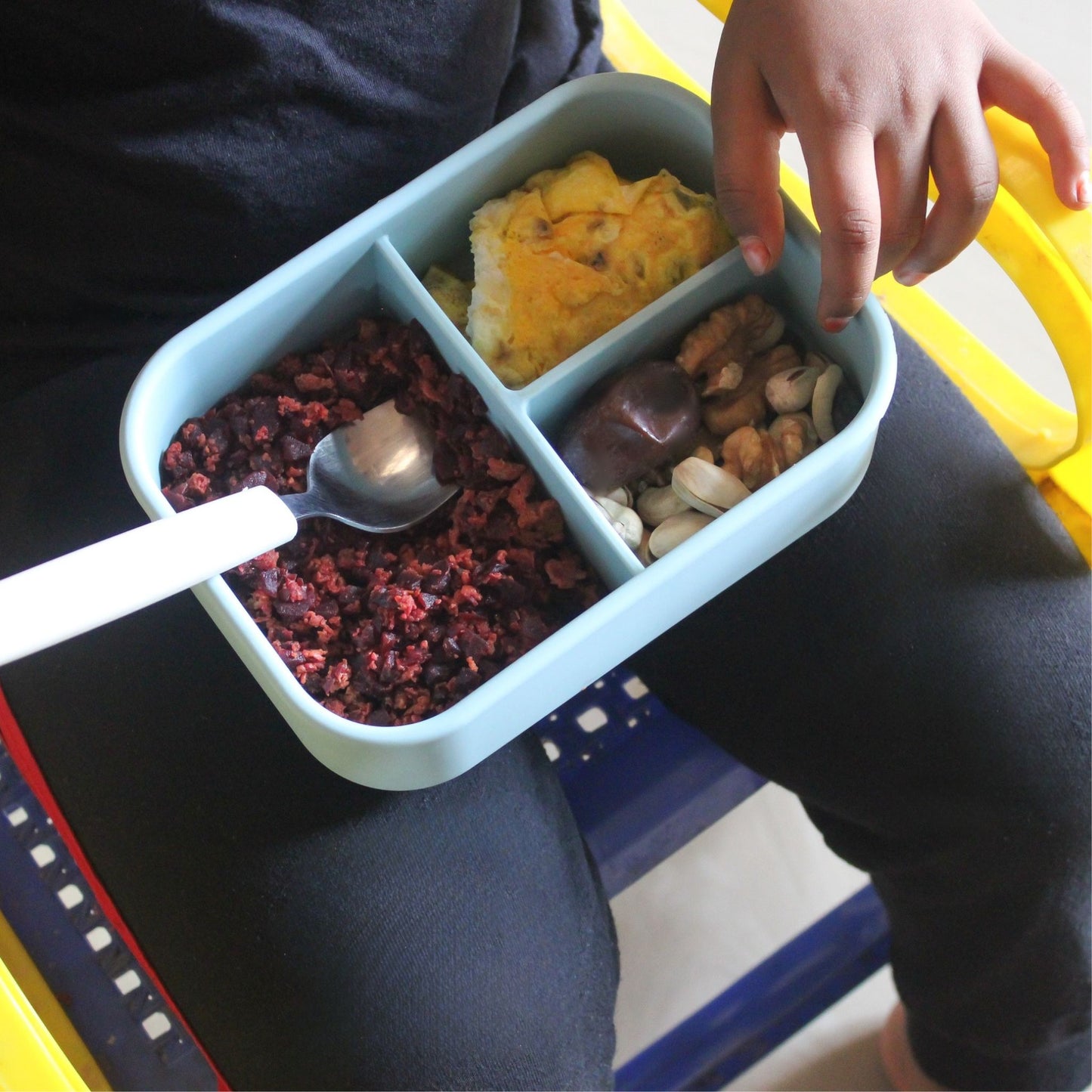 A child holding a ZipNow silicone lunchbox filled with Indian snacks—vada pav, dry fruits, and a spoonful of grated beetroot.
