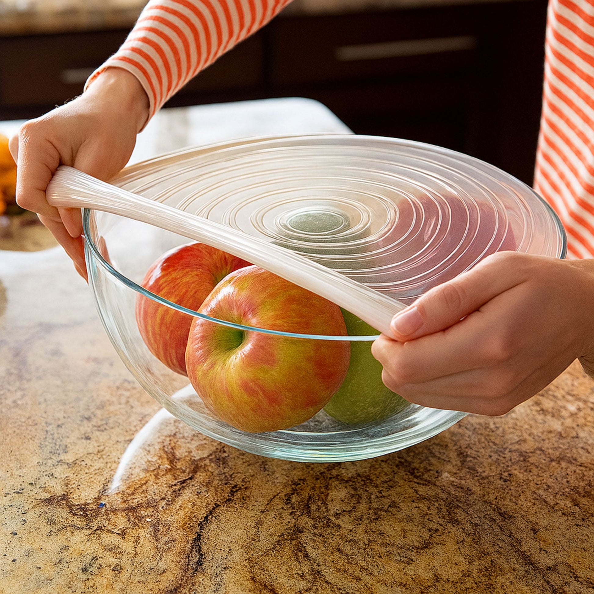 A person stretching a transparent ZipNow silicone stretch lid over a large glass bowl filled with apples, showcasing the lid’s flexibility and seal.
