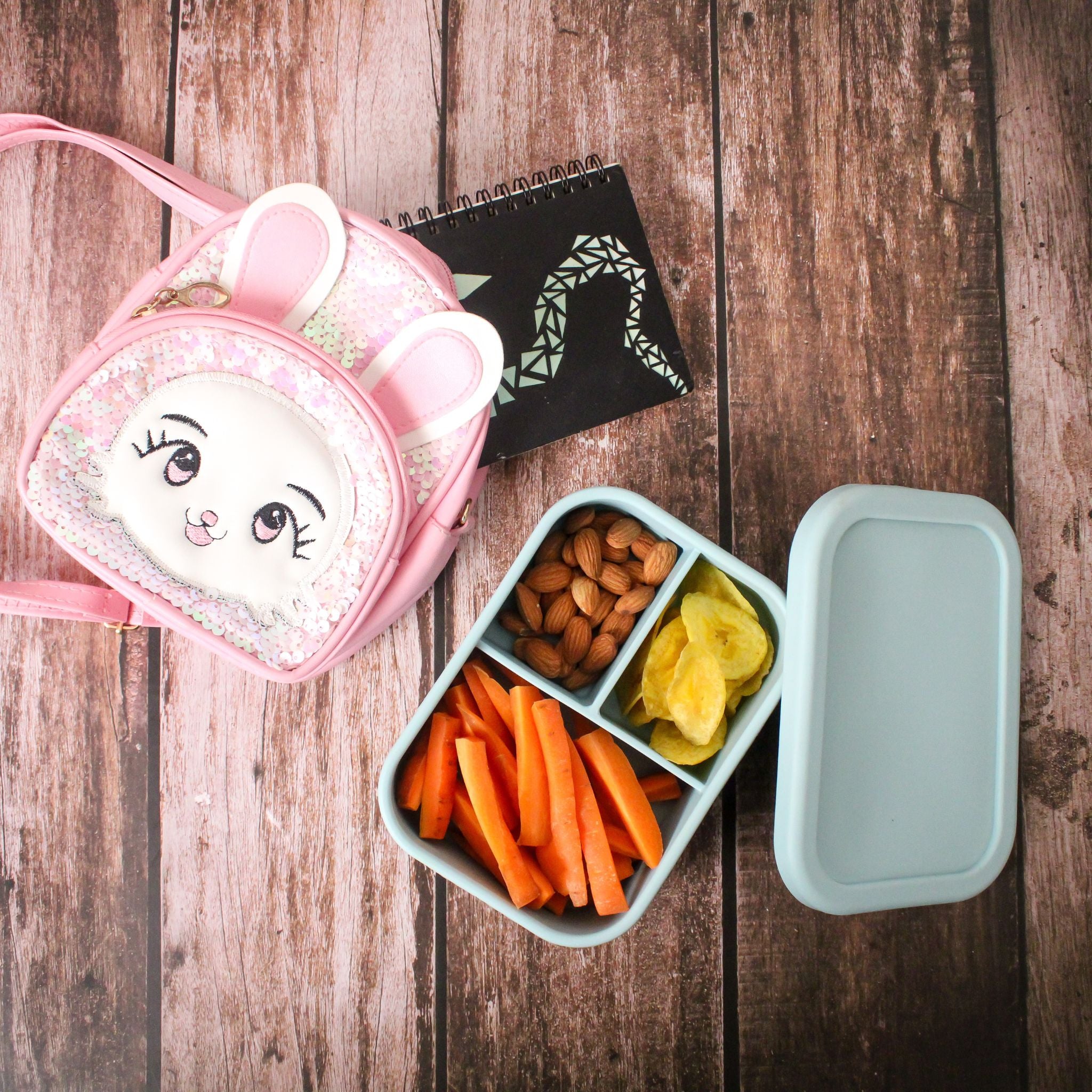 A blue ZipNow lunchbox packed with carrot sticks, almonds, and banana chips placed next to a pink kid’s backpack on a wooden background.
