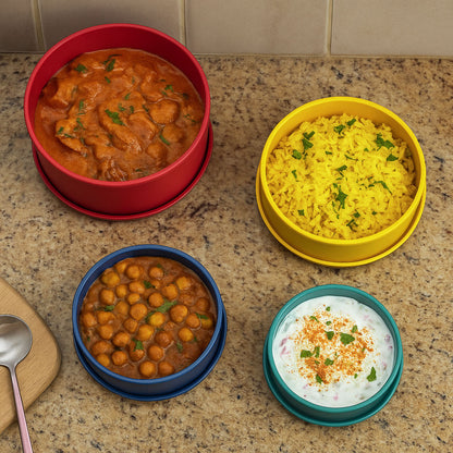 Round silicone containers in red, yellow, blue, and green filled with Indian dishes like curry, rice, and raita

