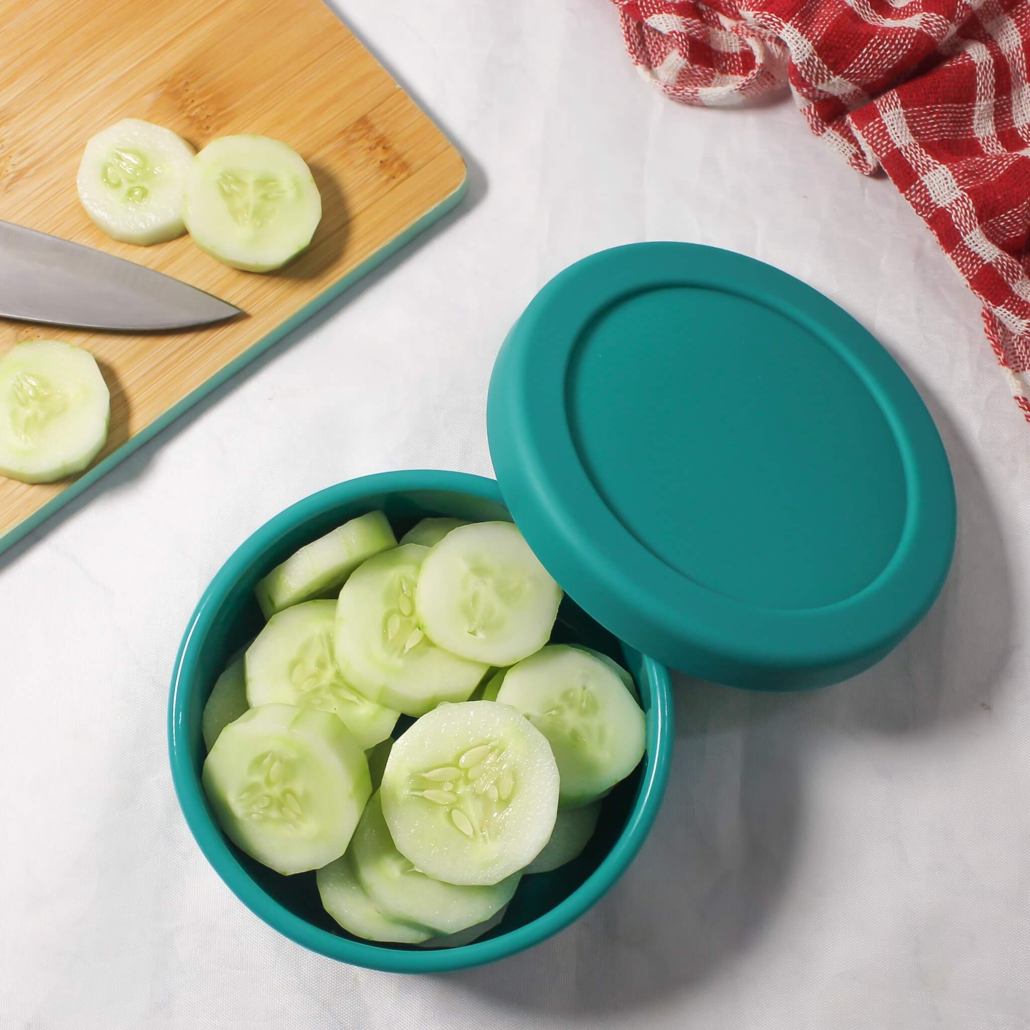 ZipNow Silicone Round container with sliced cucumbers on a white surface with a wooden cutting board and knife.